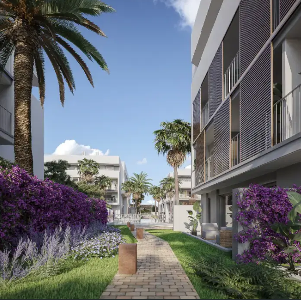 A stone-paved walkway winds through landscaped communal gardens featuring vibrant purple bougainvillea, palm trees, and modern facades with integrated sun shutters.