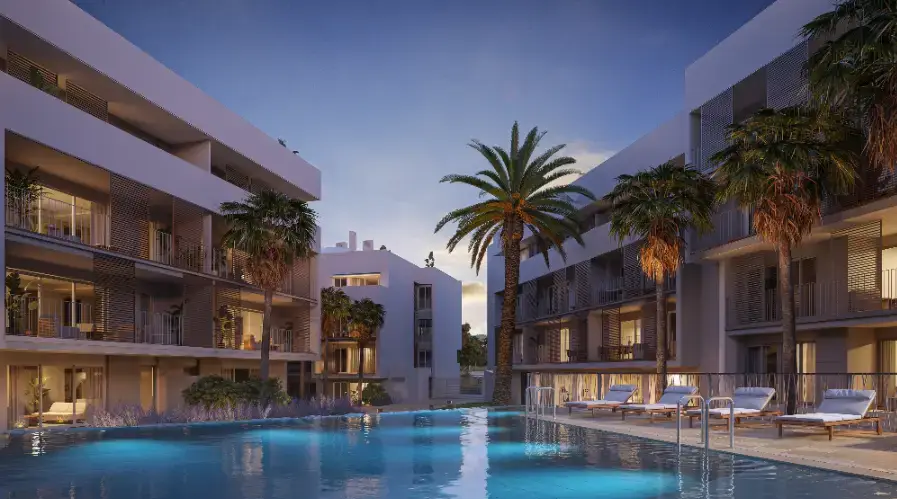 Exterior view of the communal swimming pool at dusk, featuring underwater lighting, sun loungers, and modern white facades with slatted privacy screens.