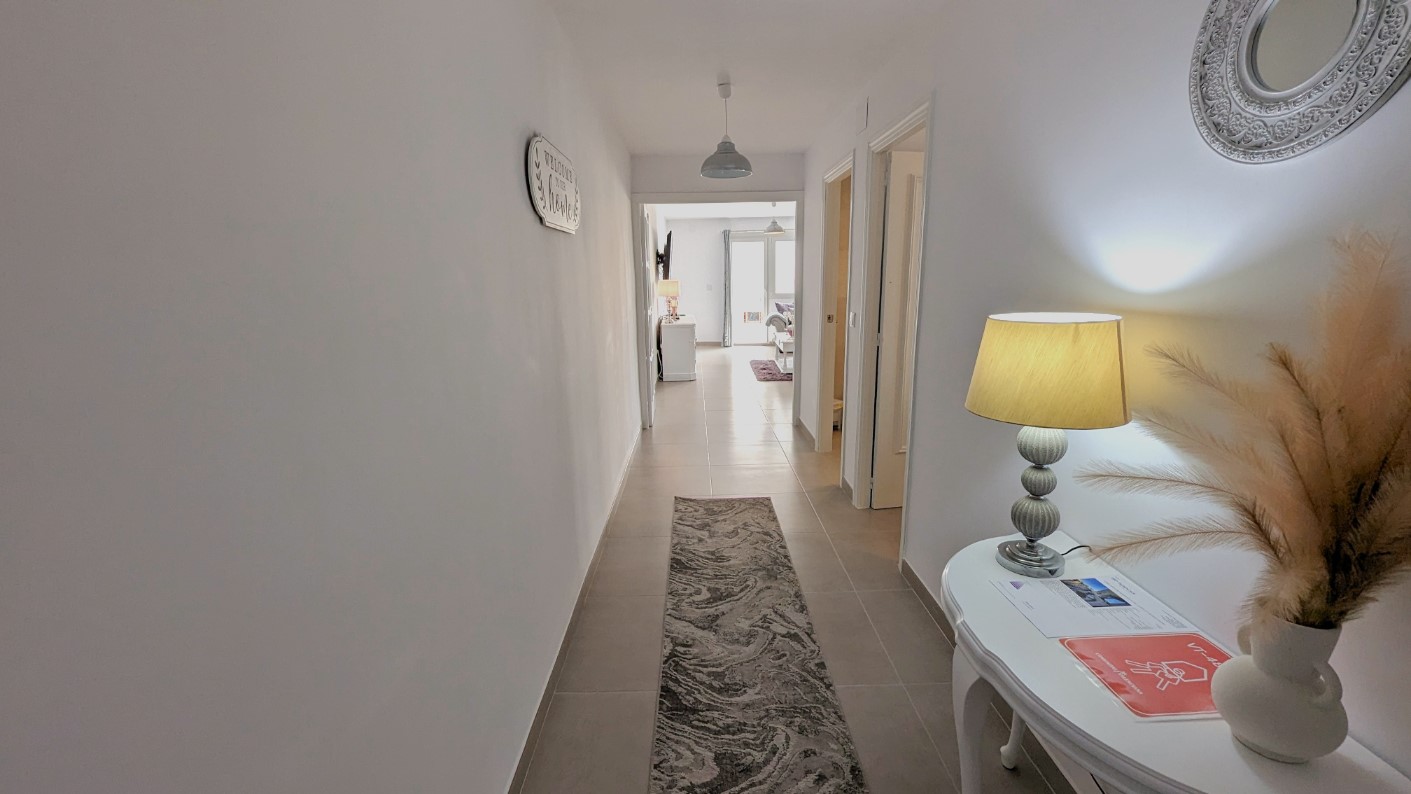 Bright entrance hallway with white walls and tiled flooring, featuring a decorative console table, ornate mirror, and a long runner leading to the living area.