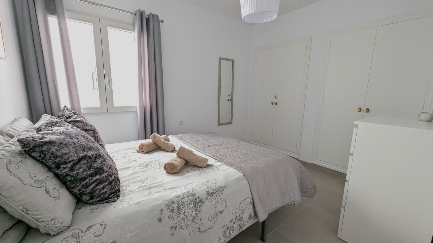 Double bedroom featuring built-in white wardrobes, a wall-mounted mirror, and tiled flooring. The space is illuminated by natural light from a window with grey curtains.