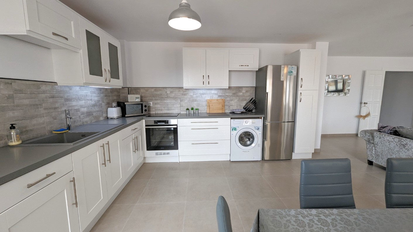 Modern open-plan kitchen featuring white shaker cabinetry, grey stone-effect backsplash, and integrated stainless steel appliances including a washing machine.
