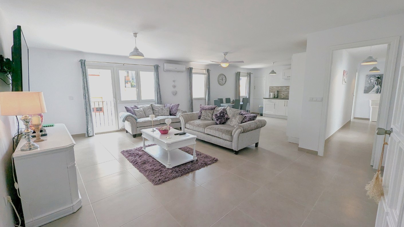 Open-plan living area featuring grey sofas with purple accents, a dining zone, and a modern white kitchen. The space includes tiled floors, a ceiling fan, and balcony access.