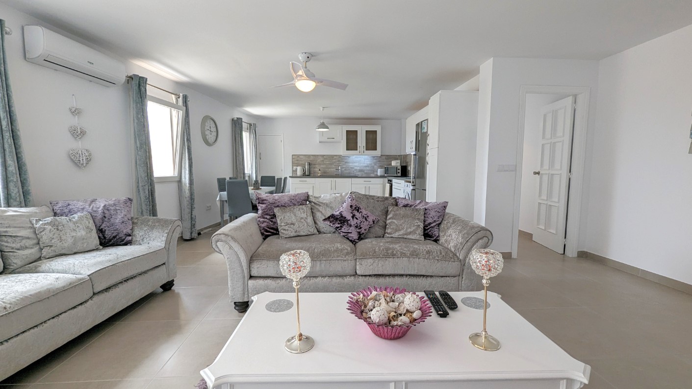 Open-plan living area featuring grey velvet sofas, a white coffee table, and a ceiling fan. The space integrates a dining area and a modern white kitchen with tiled flooring.