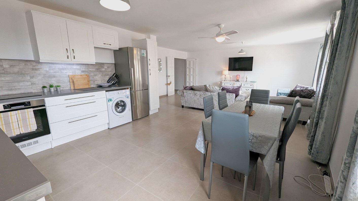 Open-plan living space featuring a modern white kitchen with integrated appliances, a central dining area with grey seating, and a lounge with a ceiling fan.