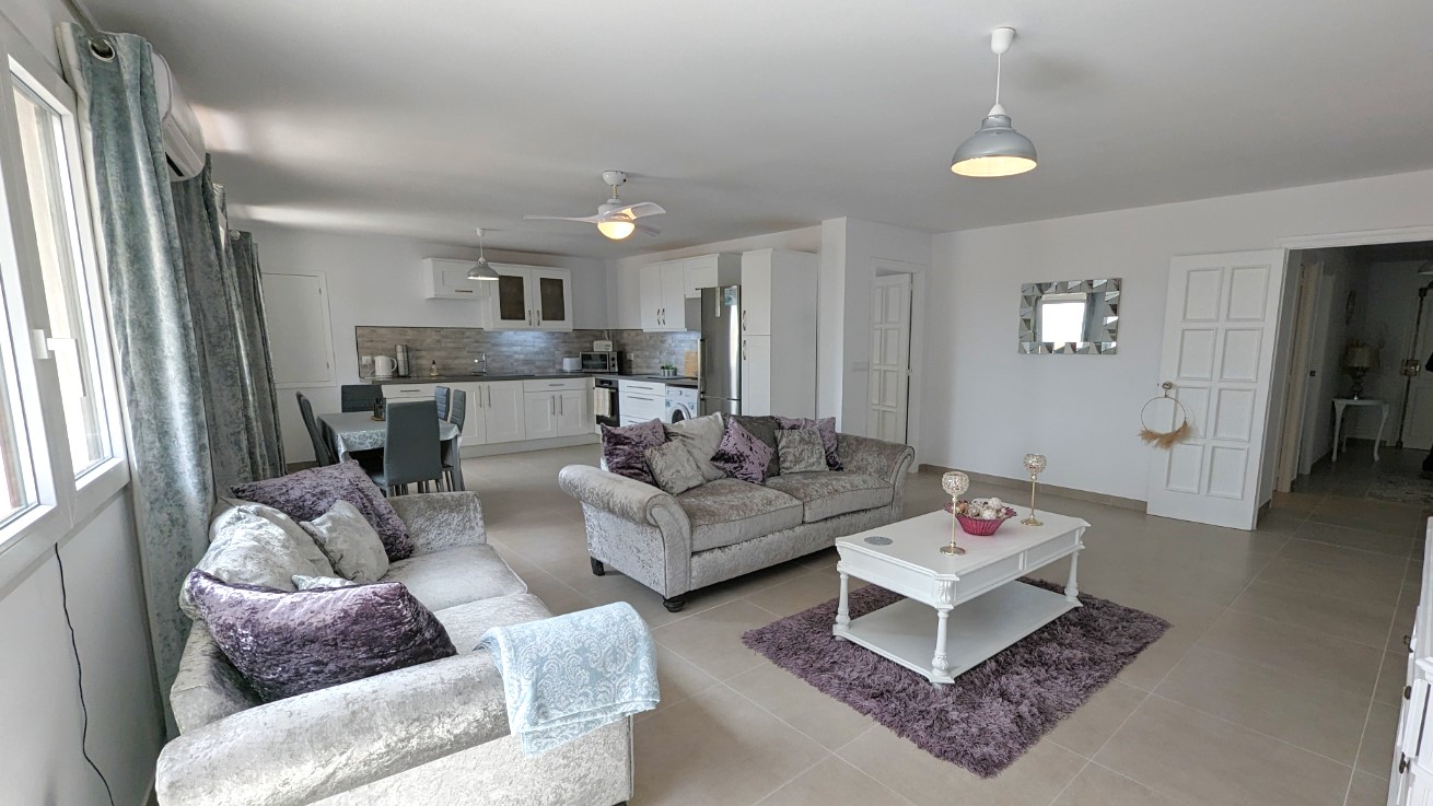 Open-plan living space featuring grey velvet sofas, a white coffee table, and a modern kitchen with white cabinetry and integrated appliances.