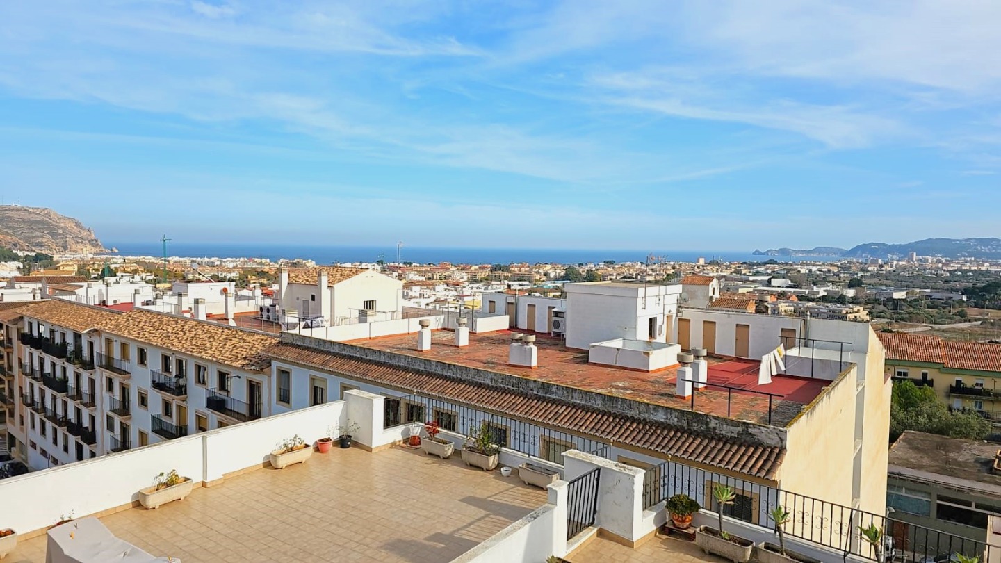 Panoramic view from the communal roof terrace overlooking Javea Old Town, the Mediterranean Sea, and distant mountains under a clear sky.