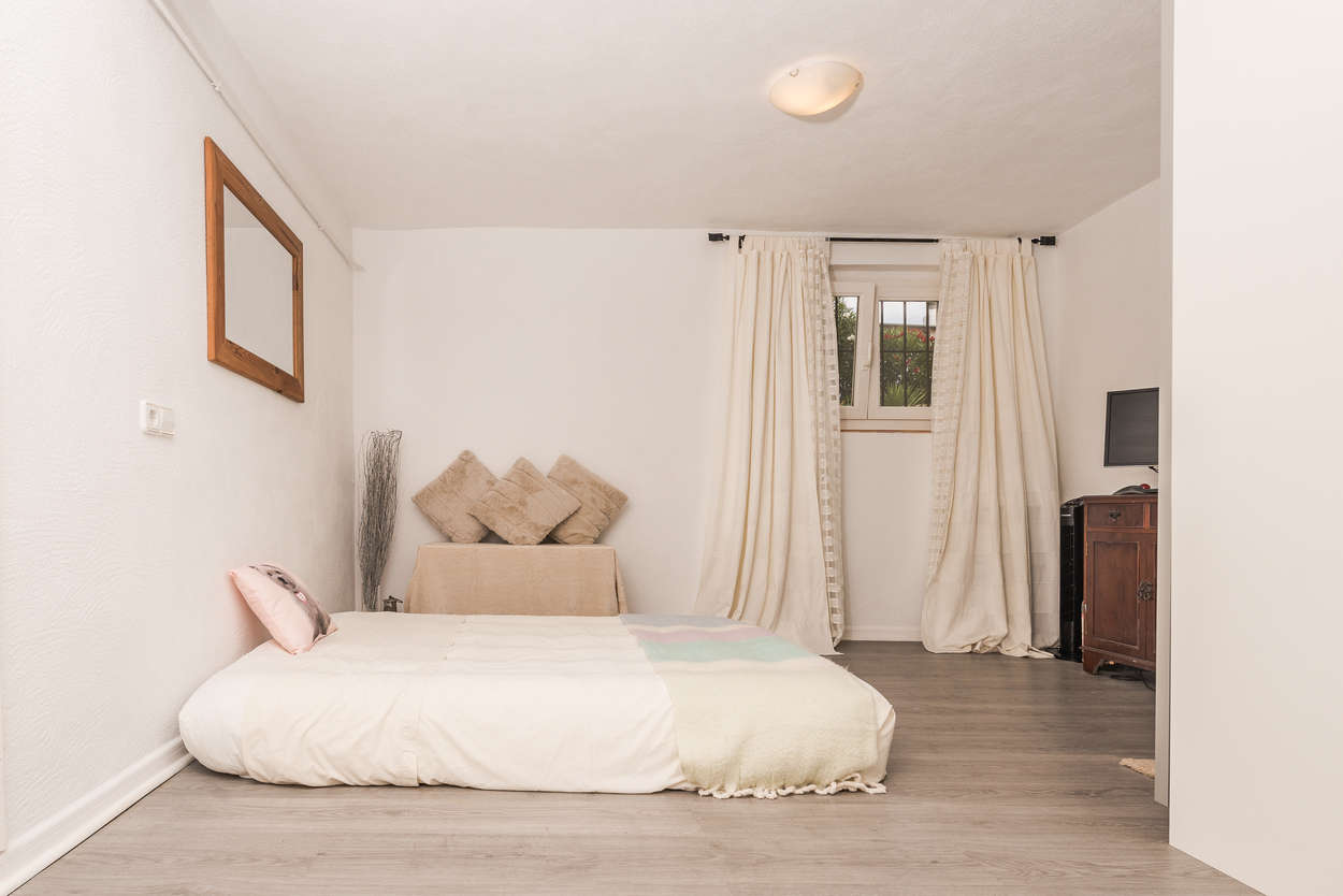 Bedroom featuring grey wood-effect flooring, white walls, and a low-profile bed. Includes a dark wood cabinet, a wooden-framed mirror, and a window with neutral curtains.