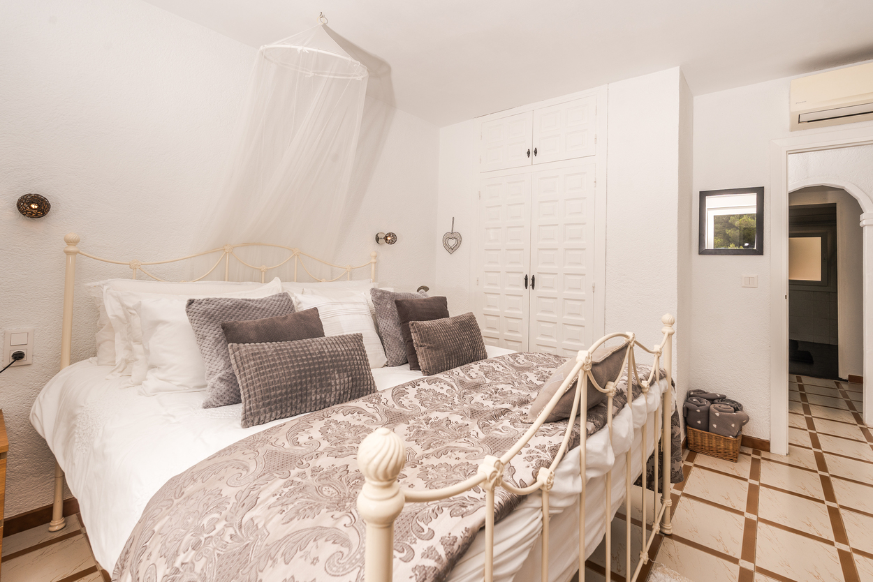 Bedroom with a white wrought-iron bed frame, mosquito net canopy, and built-in wardrobes. Features tiled flooring and an air conditioning unit above the arched doorway.