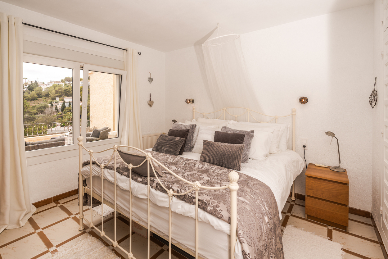 Bedroom with white metal bed frame, patterned tile floor, and double-glazed window providing natural light and views of the Mediterranean landscape.