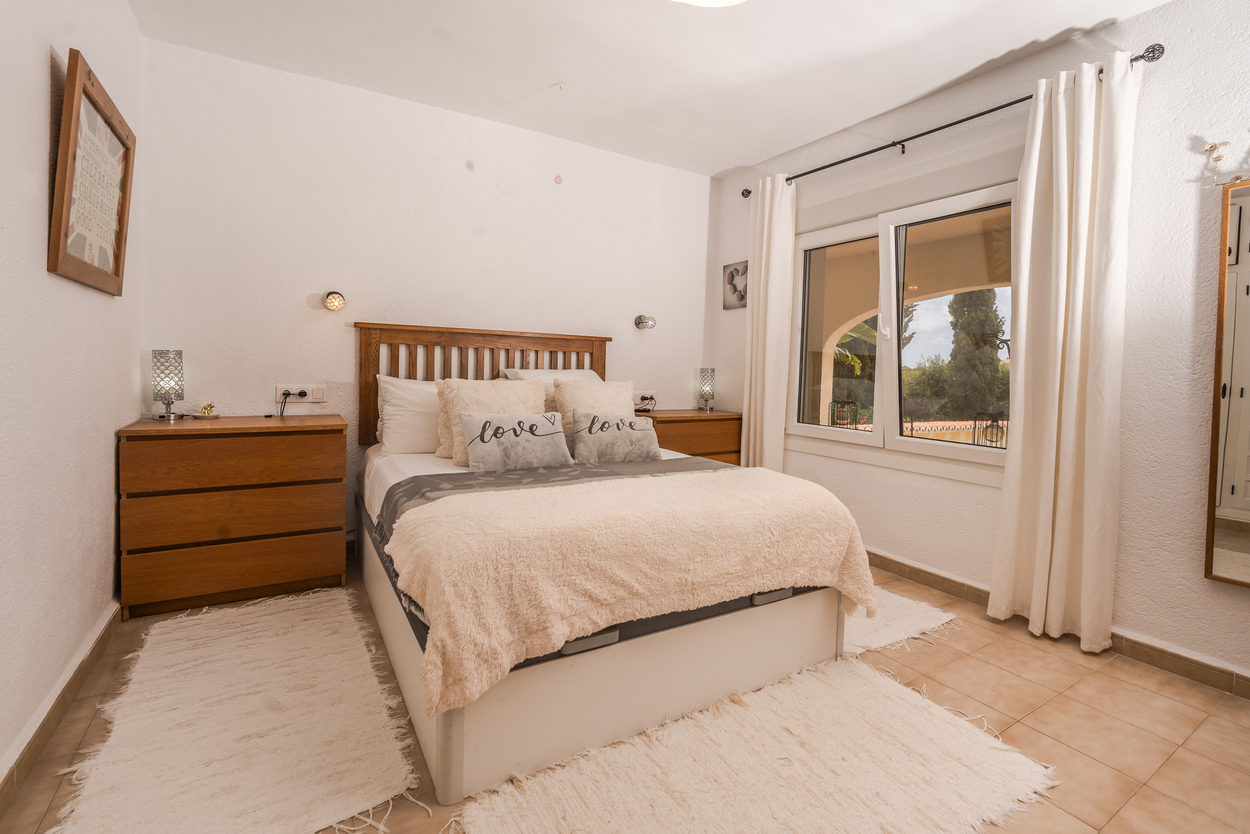 Bright bedroom featuring a wooden headboard, double-glazed windows with white curtains, and tiled flooring. Includes matching wooden chests of drawers and garden views.