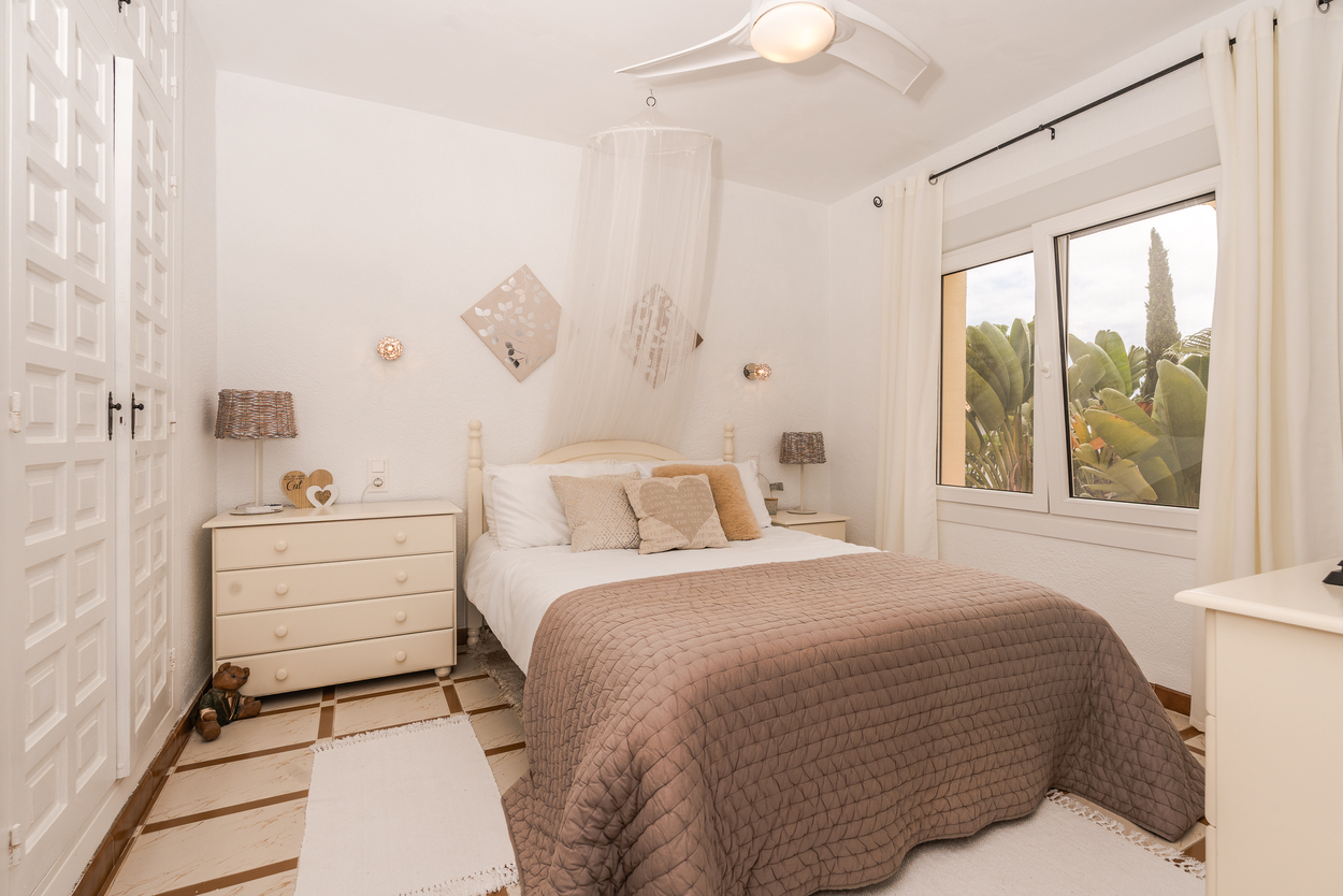 Bright bedroom featuring built-in paneled wardrobes, a ceiling fan, and double-glazed windows overlooking Mediterranean greenery with traditional tiled flooring.