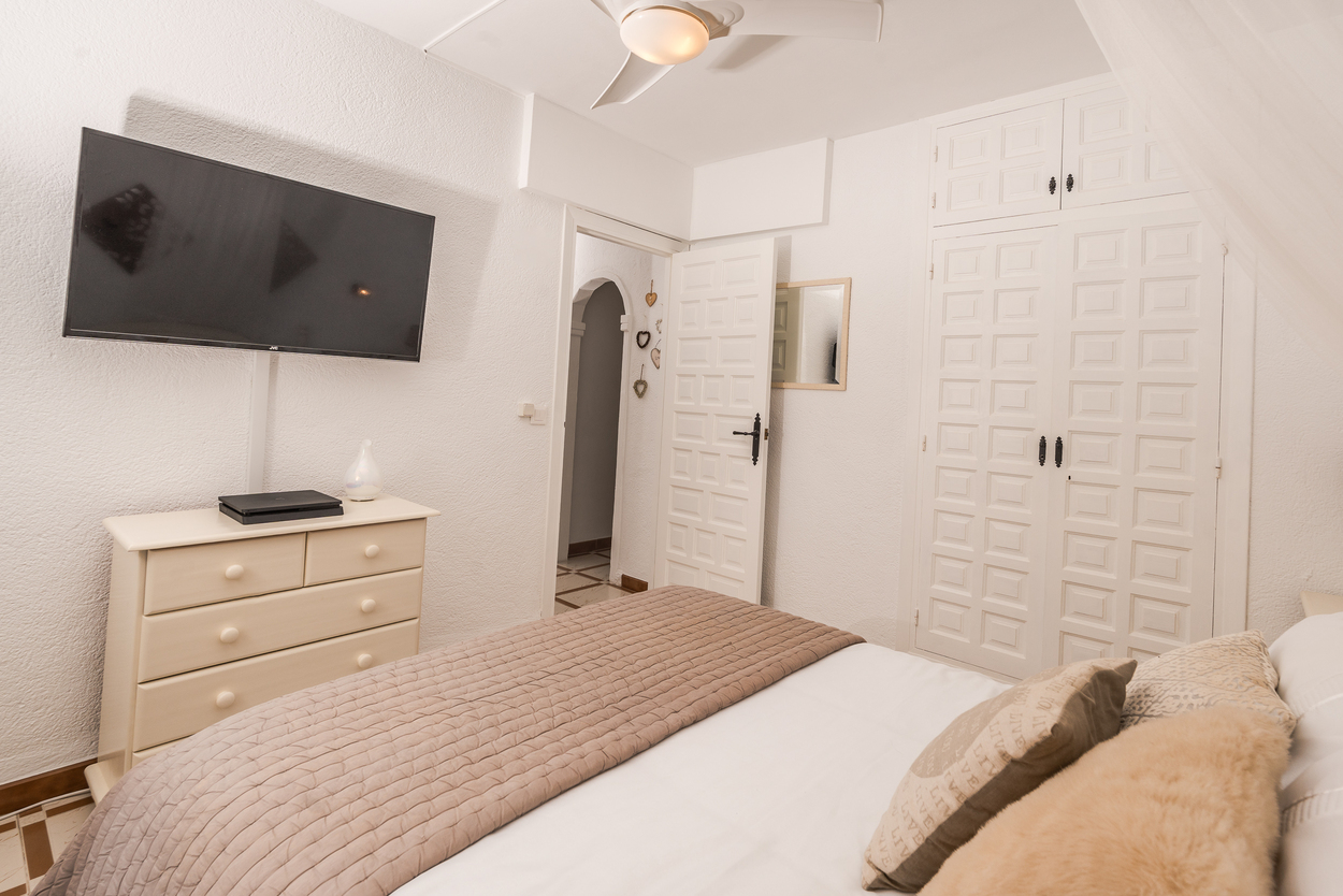 Bright bedroom featuring built-in white paneled wardrobes, a wall-mounted television, and a ceiling fan. The space includes a matching chest of drawers and tiled flooring.