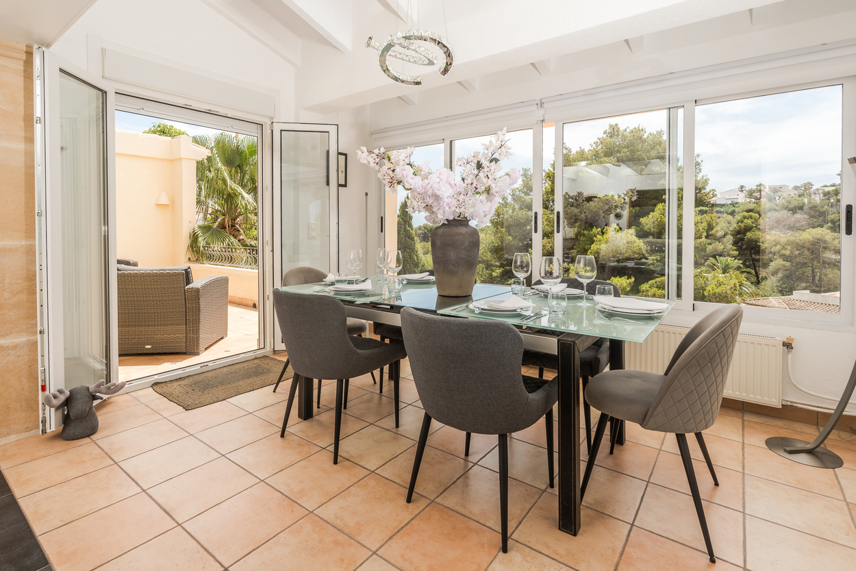 Bright dining area with terracotta floors, white beamed ceilings, and large glass doors opening onto a terrace with views of the surrounding Mediterranean greenery.