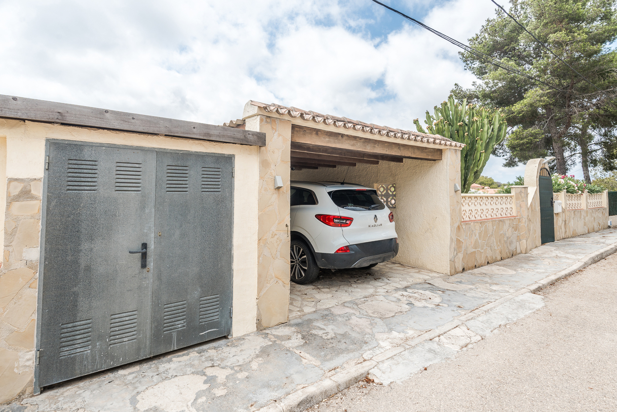 Exterior view showing the gated entrance, stone-clad walls, and a covered carport with wooden beams and a tiled roof for secure off-road parking.