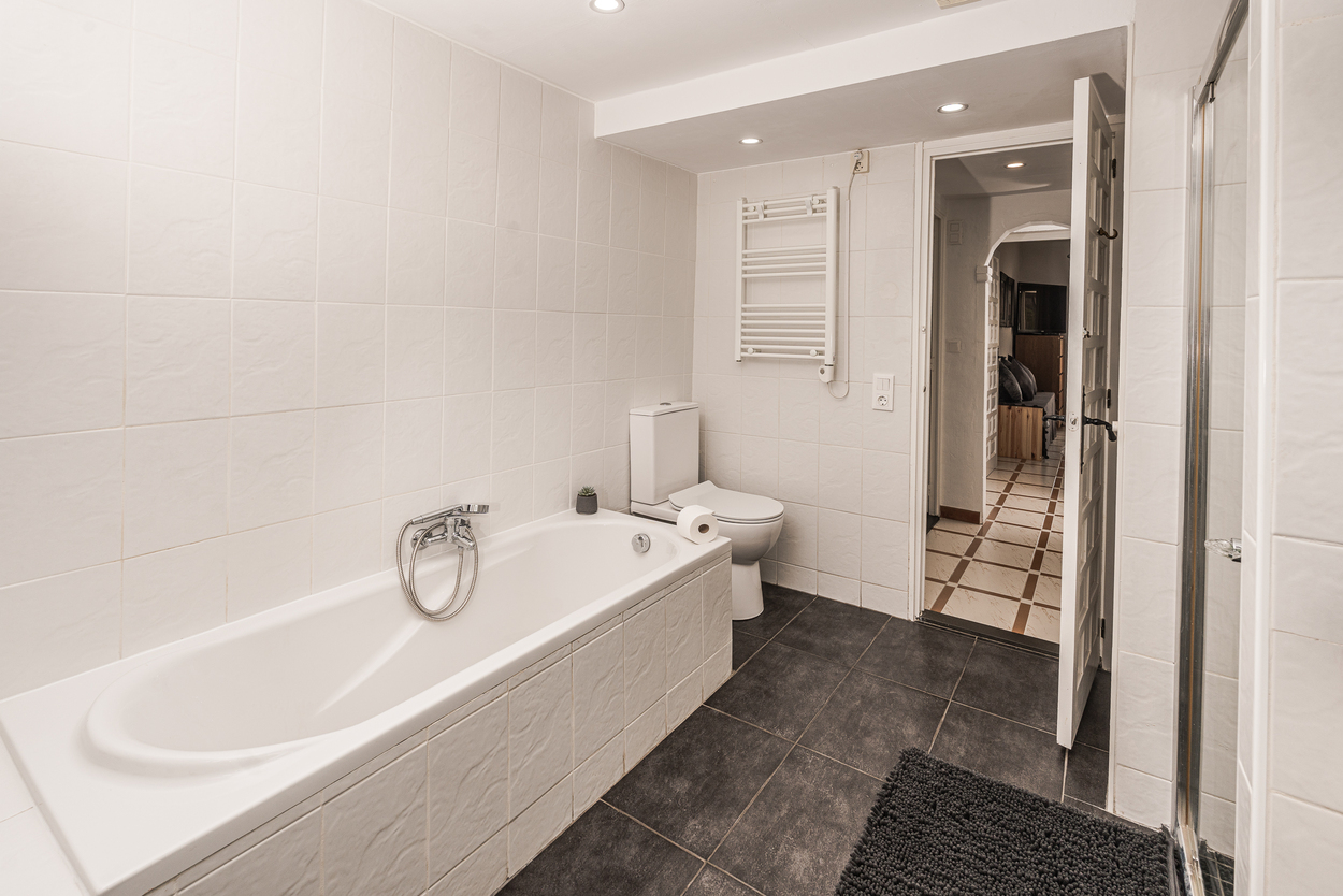 Functional bathroom featuring a white tiled bathtub, dark floor tiling, a heated towel rail, and recessed ceiling spotlights.