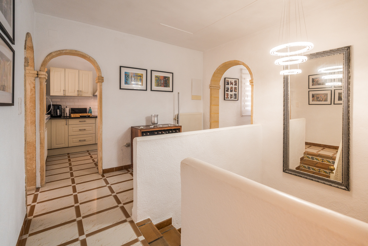 Hallway featuring traditional Tosca stone arches, tiled flooring, and a modern circular chandelier, with views into the kitchen and a decorative staircase.