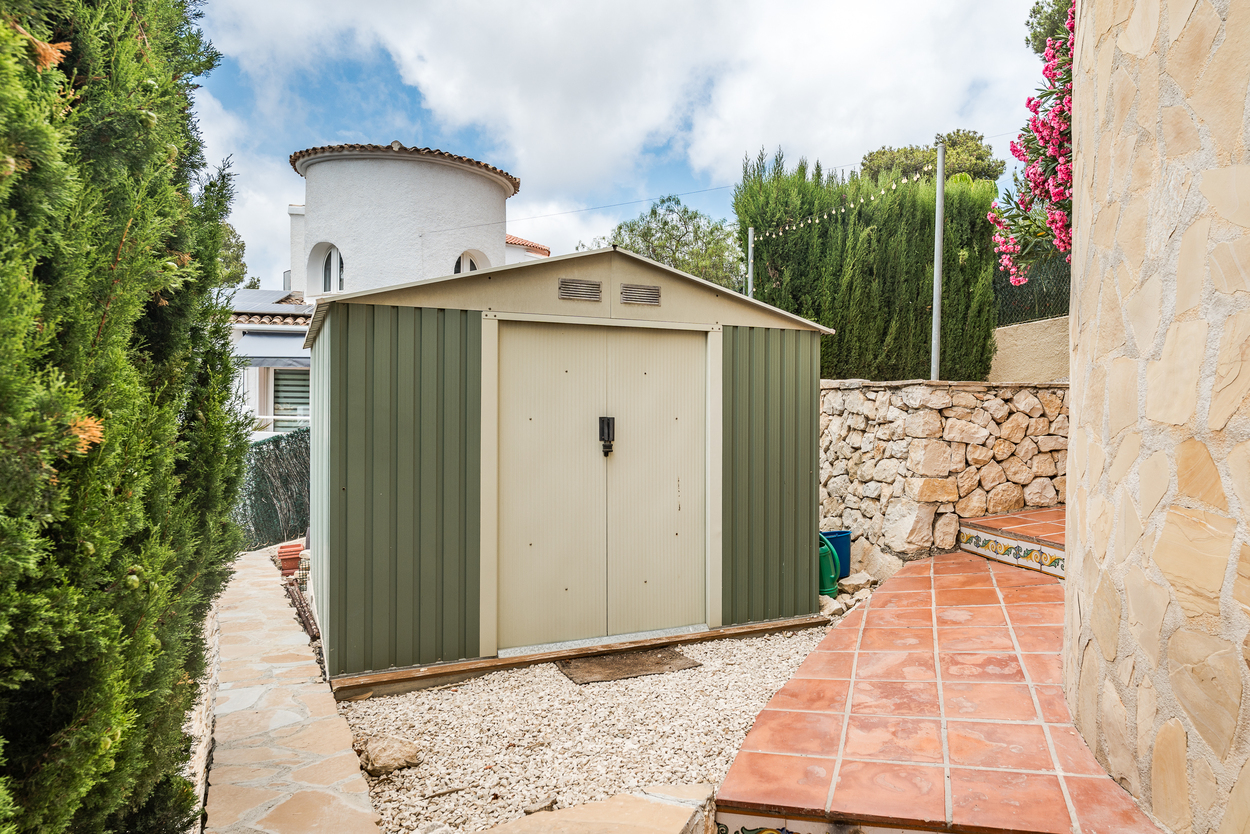 Metal garden storage shed situated on a gravel area, bordered by stone walls, tiled steps, and mature privacy hedging.