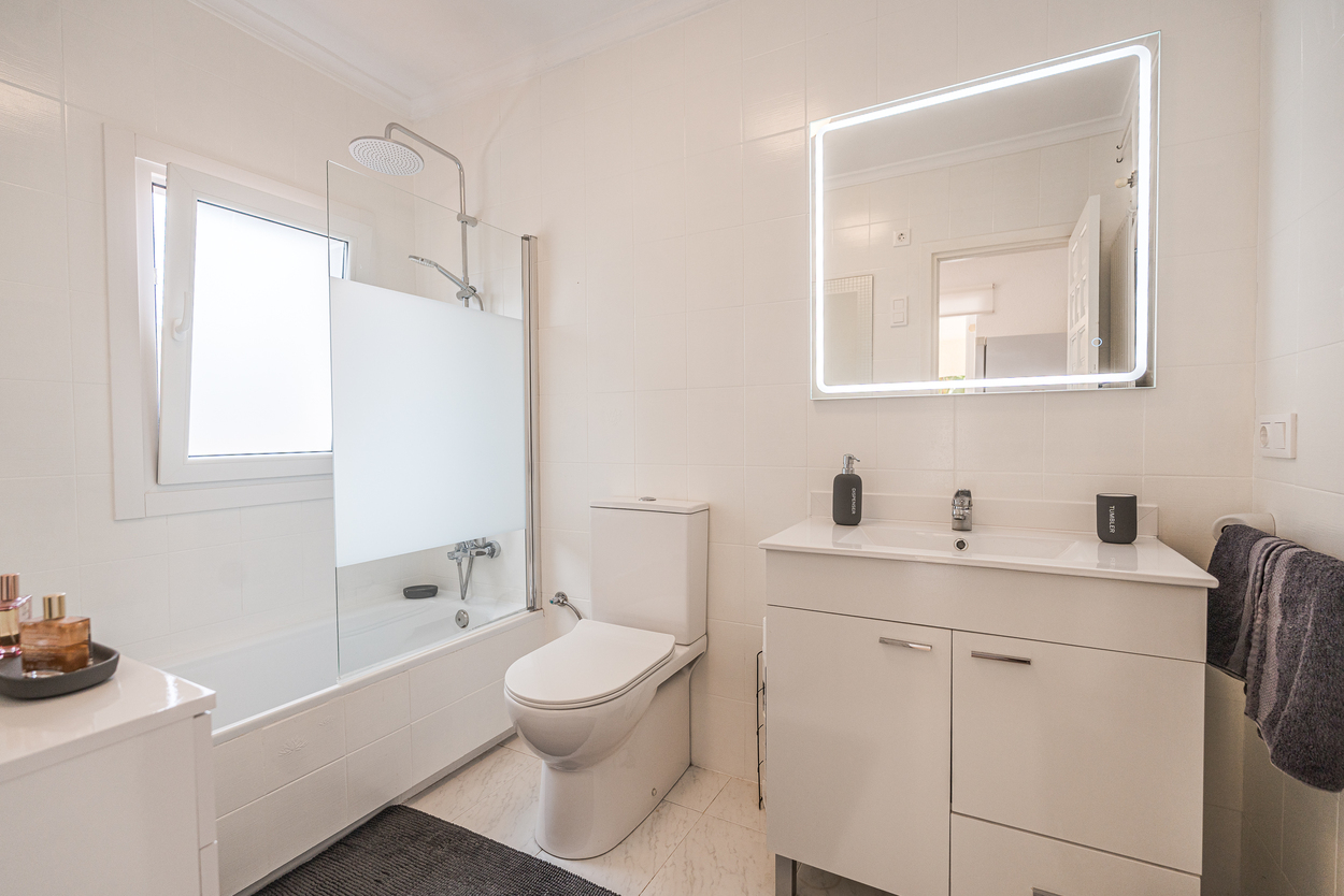 Modern bathroom featuring white tiling, a vanity with an LED-backlit mirror, and a bathtub equipped with a glass screen and rainfall shower head.
