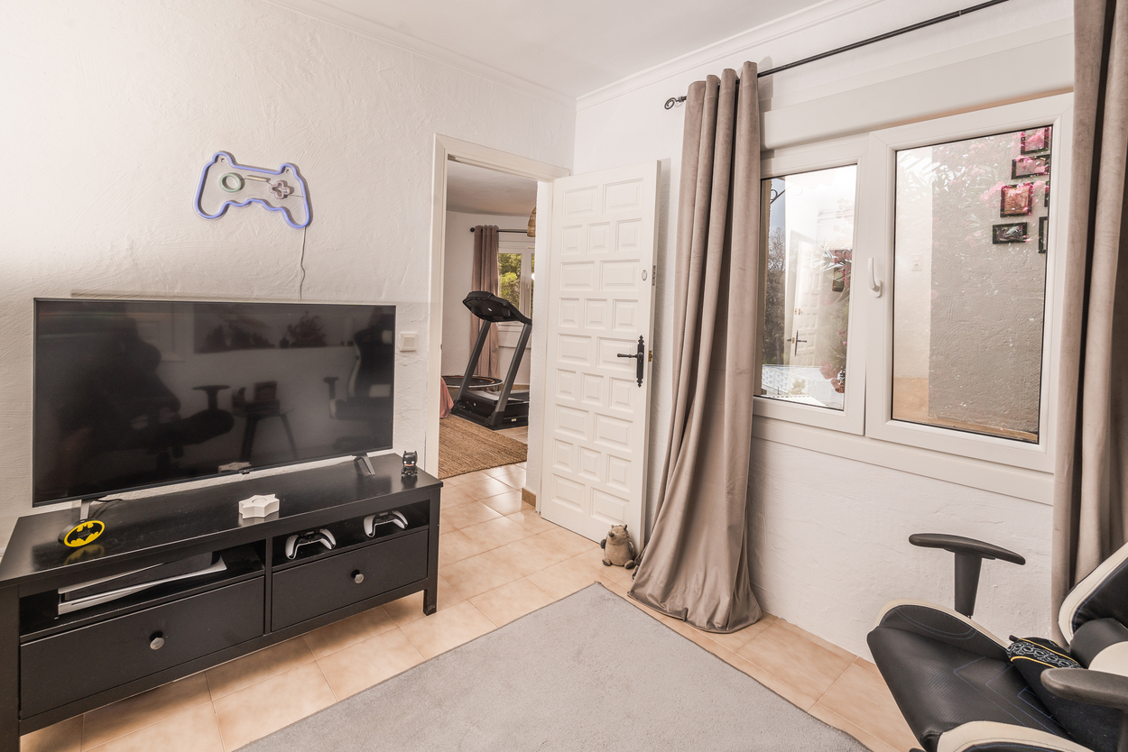 Multimedia room with gaming setup, flat-screen TV, and tiled flooring, offering a view into an adjacent fitness area with a treadmill.