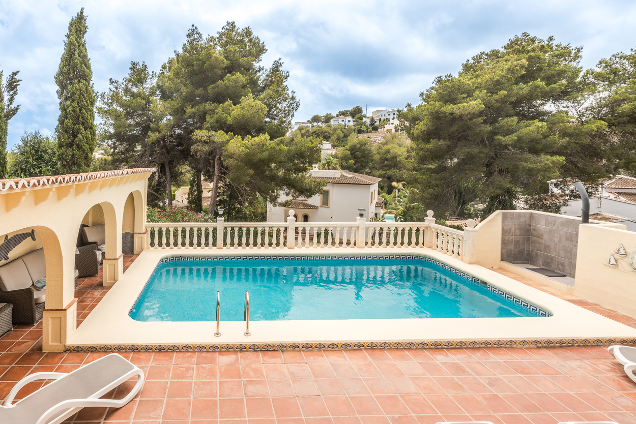 Outdoor terrace featuring an 8x4m pool with mosaic tiling, a white balustrade, and a covered arched naya with seating, surrounded by mature pine trees.