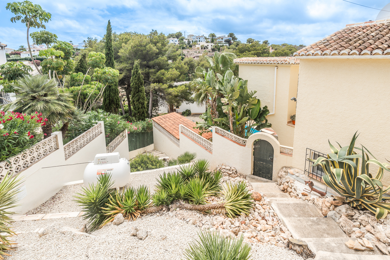 Terraced Mediterranean garden with white gravel, yuccas, and agaves. Concrete steps lead toward a gated arched entrance and the villa's traditional tiled roofs.