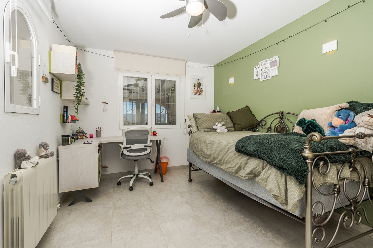 Bedroom featuring a green accent wall, an ornate metal bed frame, and a functional workspace with a built-in desk and ergonomic chair. Includes a ceiling fan and radiator.