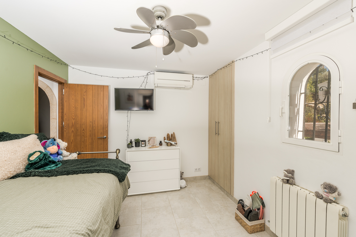 Bright bedroom featuring a green accent wall, built-in wardrobes, and an arched window. Includes a ceiling fan, air conditioning, and a wall-mounted television.