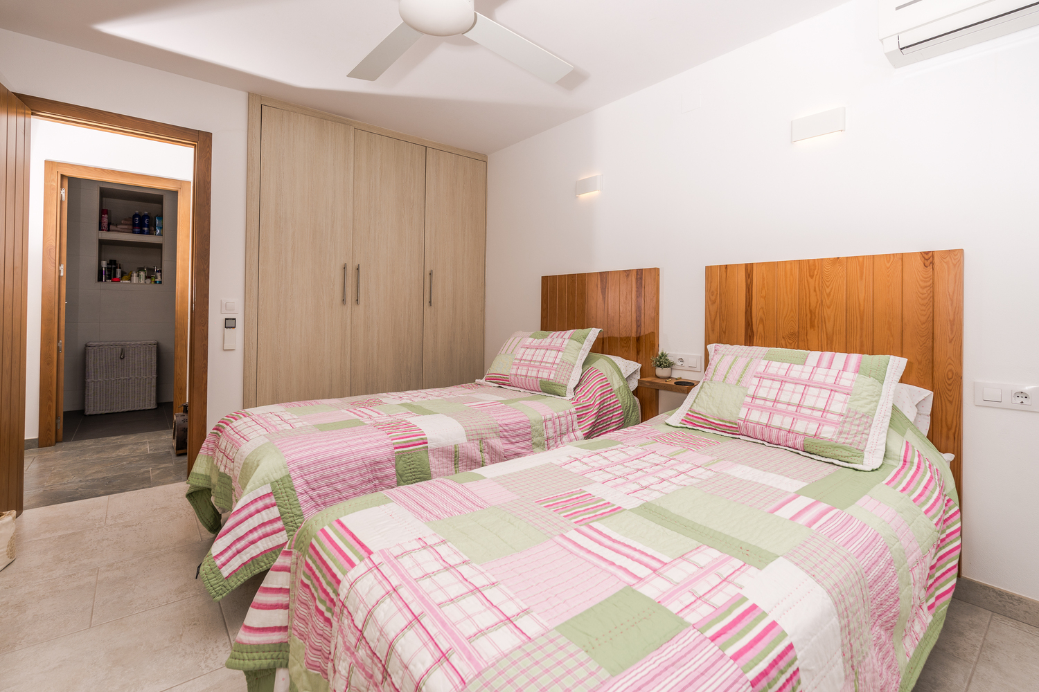 Bright bedroom featuring twin beds with natural wood headboards, built-in wardrobes, a ceiling fan, and access to an en-suite bathroom.