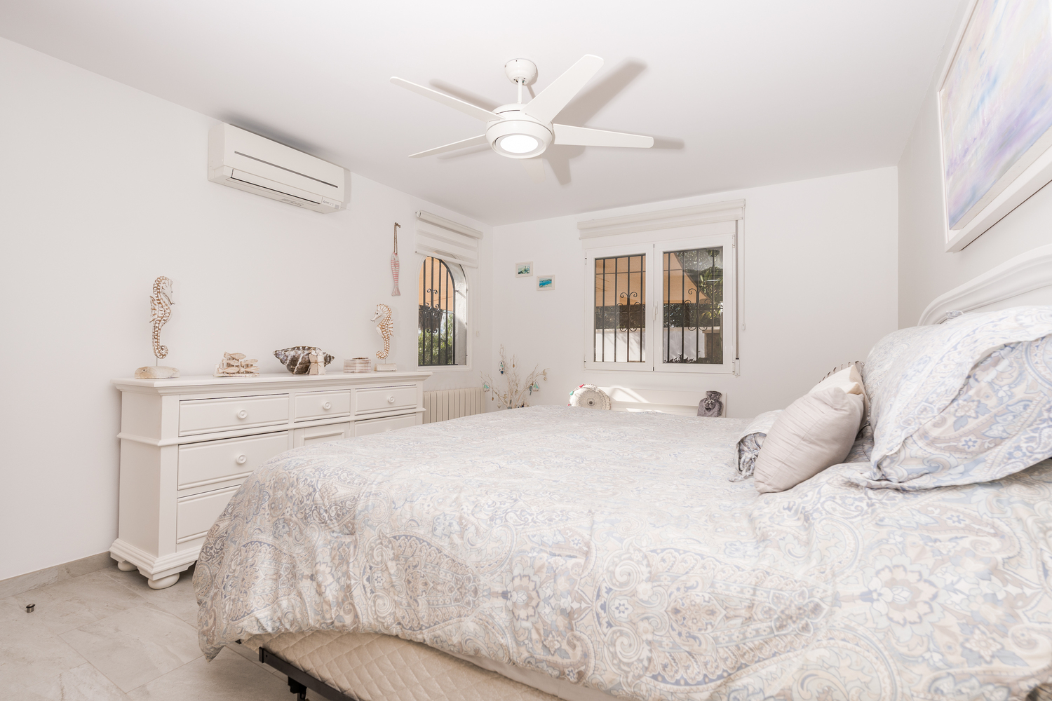 Bright bedroom featuring white walls, a modern ceiling fan, air conditioning, and a classic white dresser. Large windows with security bars provide natural light.