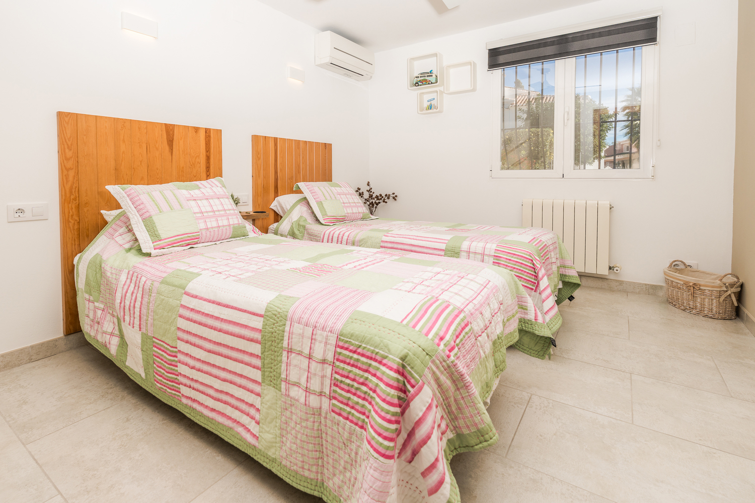 Bright twin bedroom featuring wooden headboards, tiled flooring, and split-system air conditioning. A large window with security bars provides natural light.