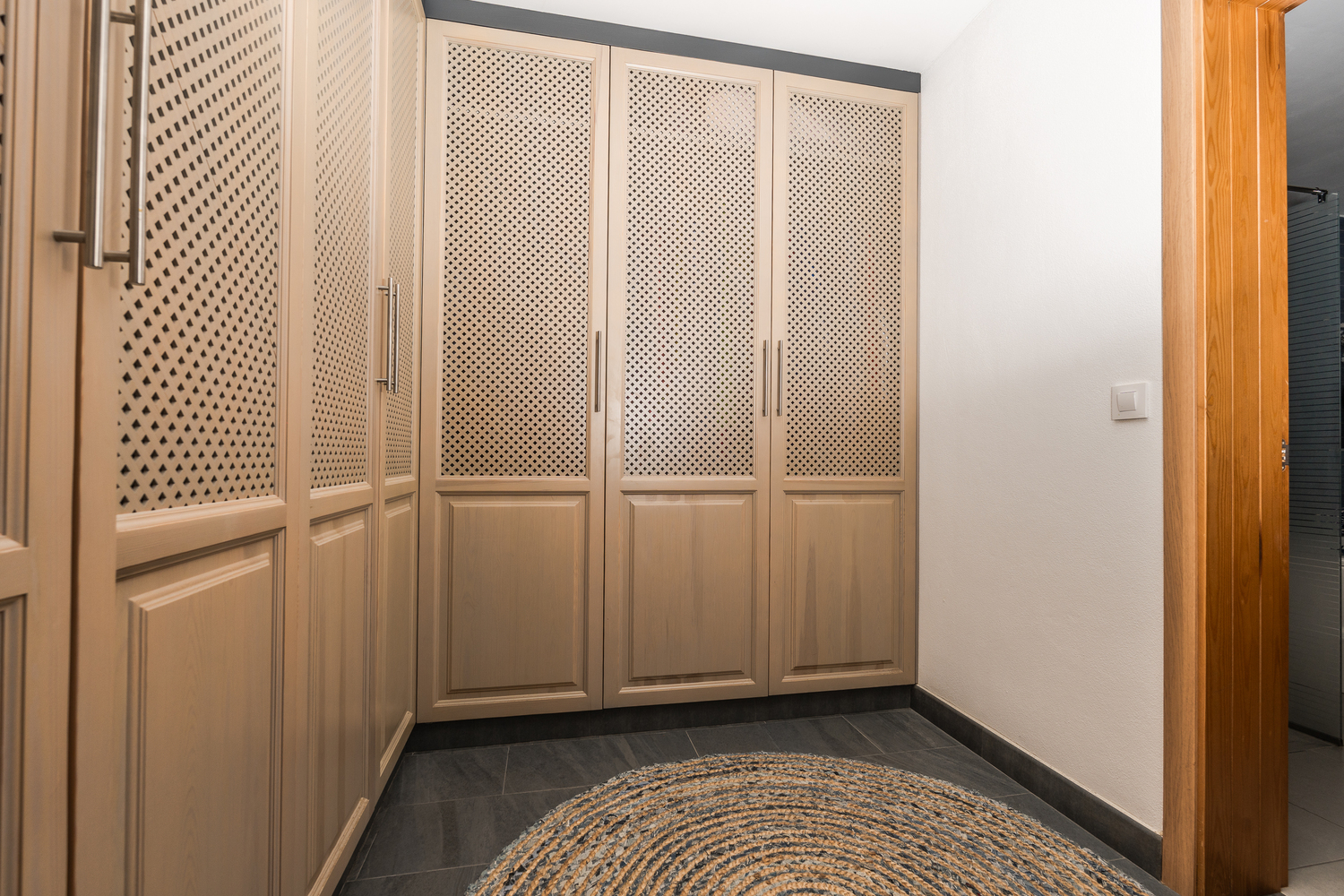 Dressing room featuring floor-to-ceiling wooden wardrobes with decorative lattice panels, dark tiled flooring, and a circular woven rug.
