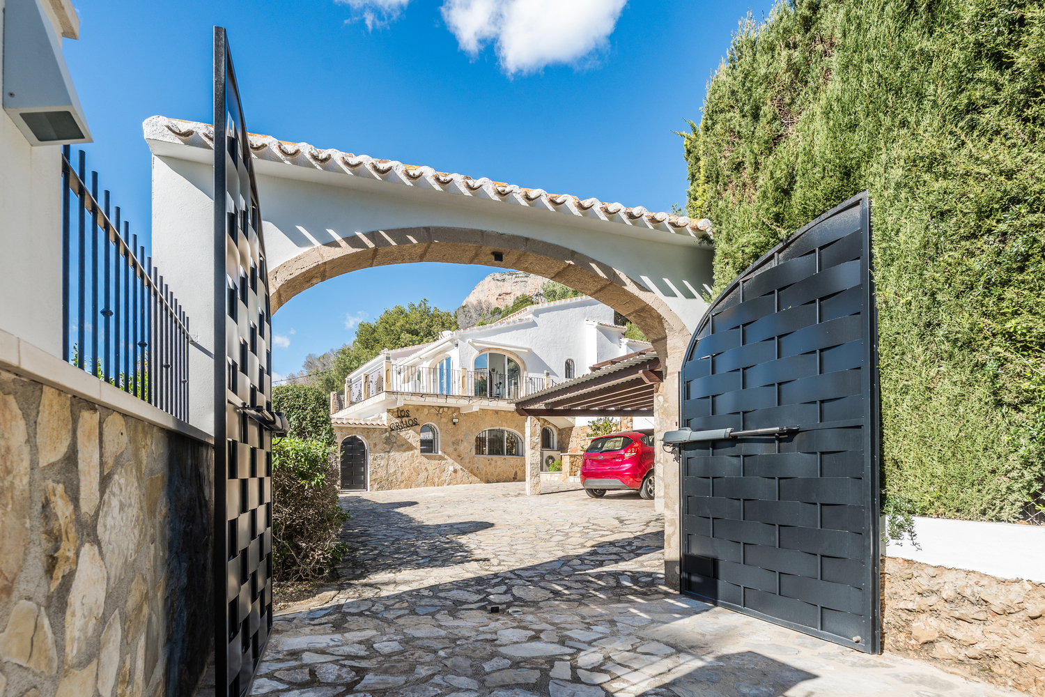 Entrance featuring automated black metal gates and a traditional Tosca stone arch, opening onto a stone-paved driveway with a view of the villa and carport.