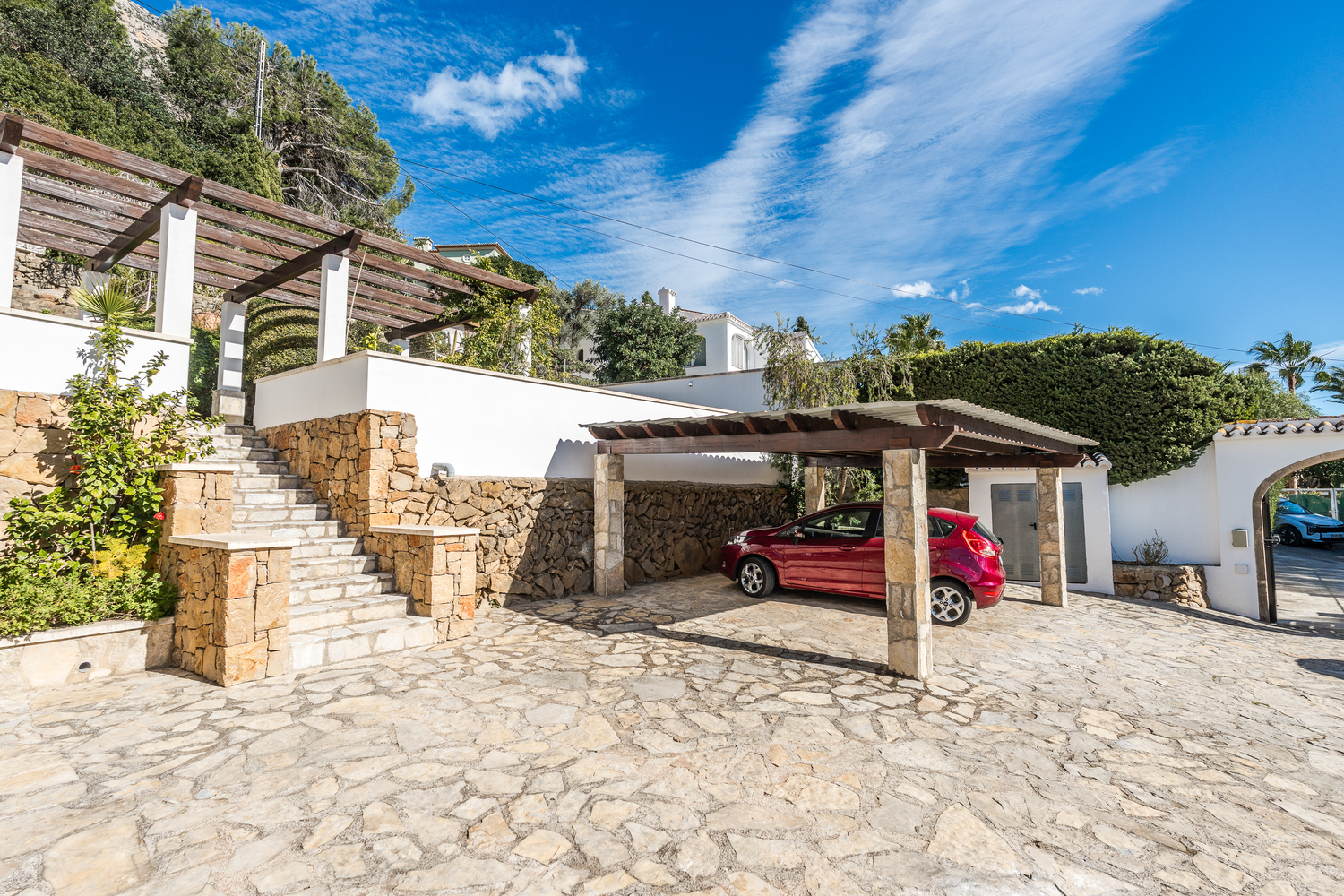Exterior entrance featuring a stone-paved driveway, a covered carport with timber beams, and natural stone masonry walls leading to the upper terraces.