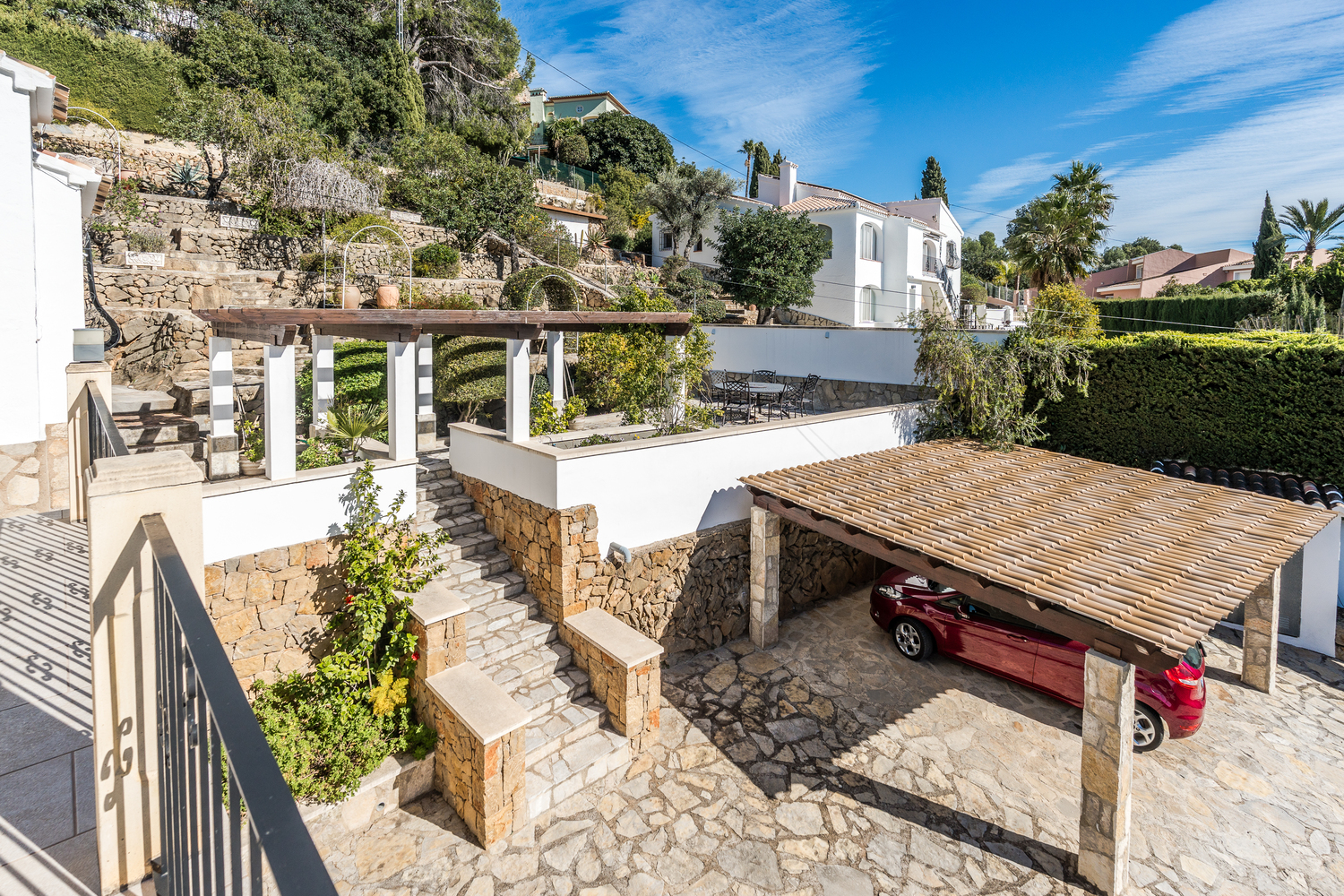 Exterior view featuring a stone-paved driveway, a covered carport with a tiled roof, and terraced Mediterranean gardens connected by stone stairs.