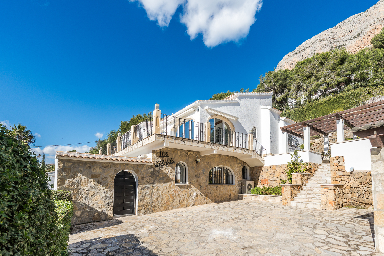 Exterior view featuring the natural stone facade, arched windows, and a spacious stone-paved driveway set against the Montgó mountain.