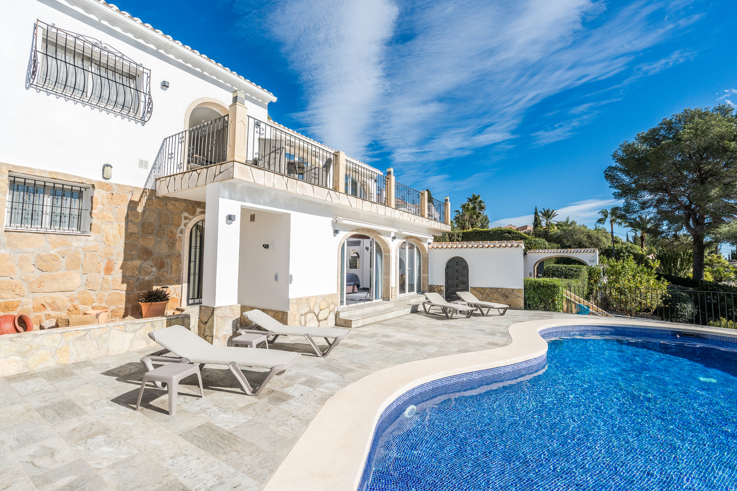 Exterior view of the curved mosaic pool and stone terrace, featuring white sun loungers and the villa's white facade with natural stone accents and arched doorways.
