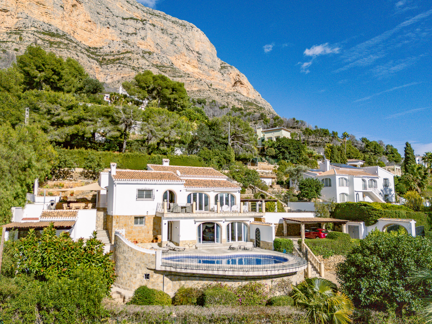 Exterior view of the two-level villa at the foot of the Montgó mountain, featuring a curved swimming pool, white facades with stone cladding, and arched terrace windows.