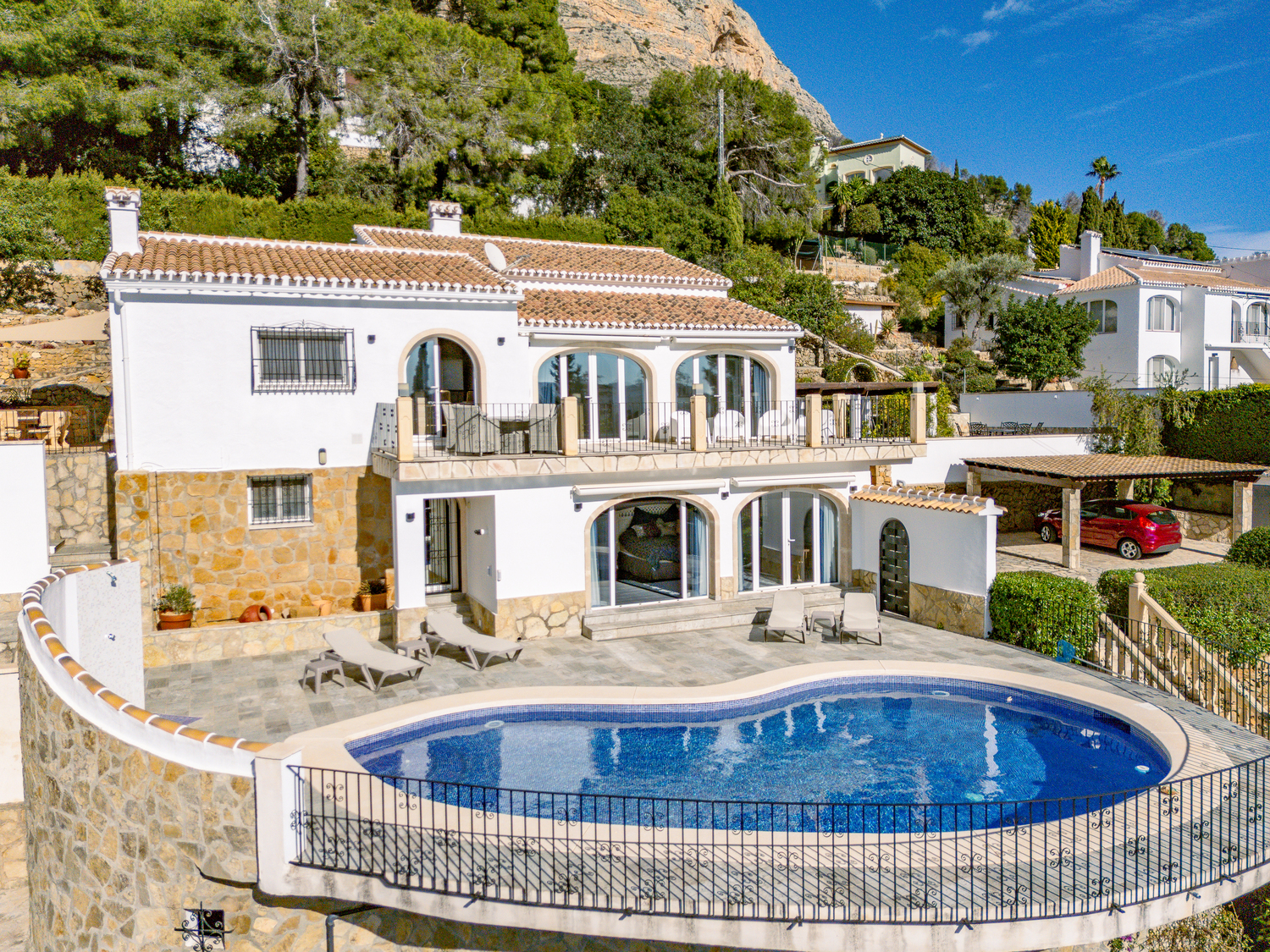 Exterior view of the two-level villa featuring a curved swimming pool, stone-paved terrace, and arched glass doors set against the Montgó mountain backdrop.