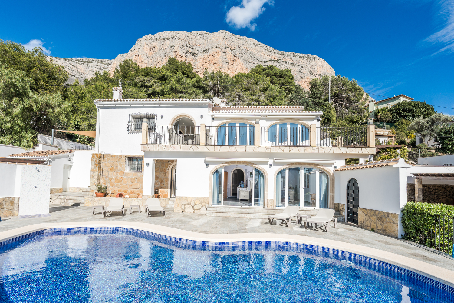 Exterior view of the two-story villa featuring a curved swimming pool, stone-paved terrace, and arched glass doors against the Montgó mountain backdrop.