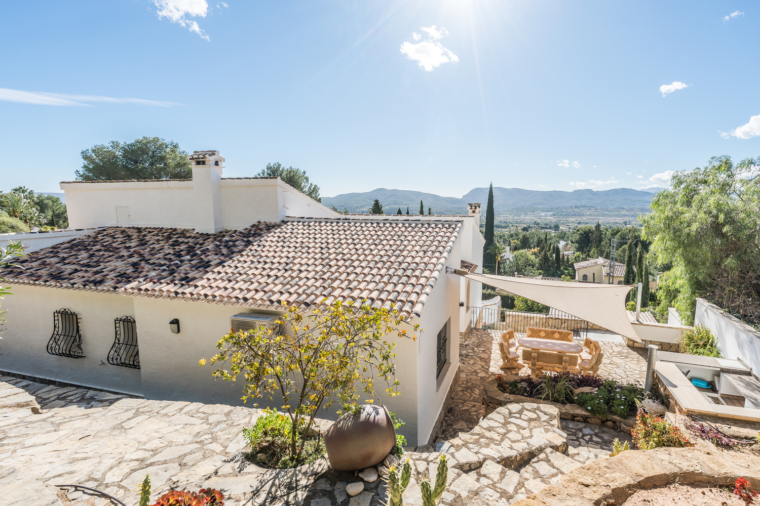Exterior view of the villa featuring traditional terracotta roofing, stone-paved terraces, a shaded dining area with a sun sail, and panoramic mountain views.