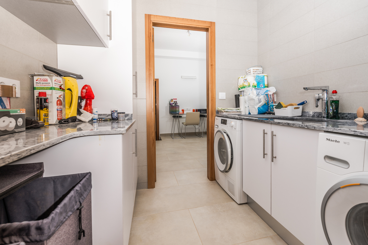 Functional laundry room featuring white cabinetry, grey marble countertops, and high-end Miele appliances, with a view into the adjacent home office space.