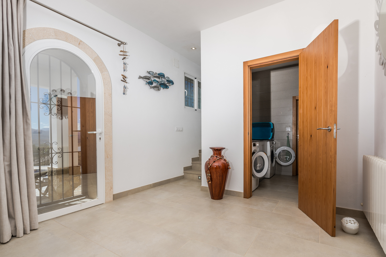 Hallway with tiled floors featuring an arched glass door with stone trim, a staircase, and a wooden door opening to a laundry room with appliances.