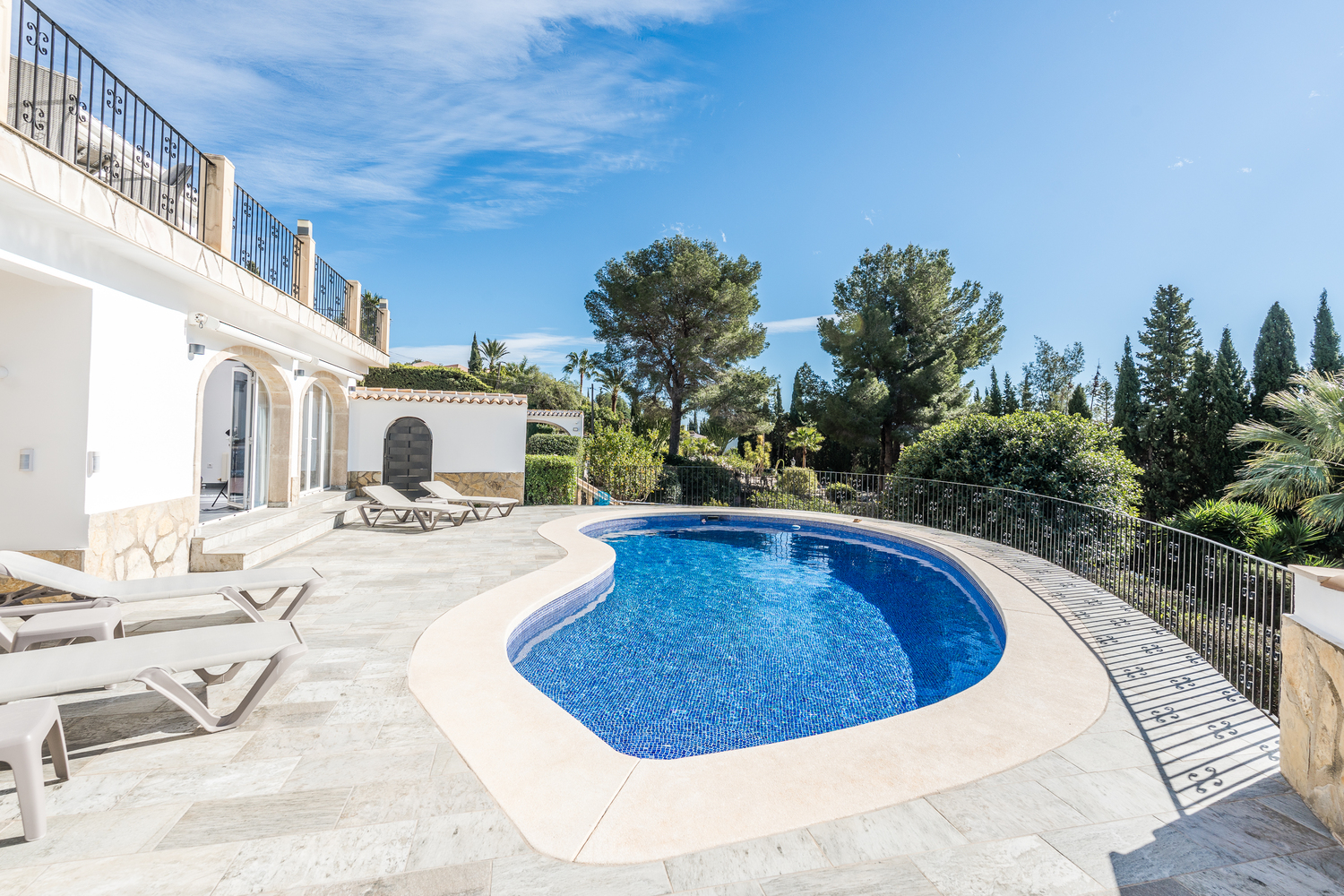 Kidney-shaped swimming pool surrounded by a light stone terrace with sun loungers, set against a backdrop of mature Mediterranean trees and a white villa facade.