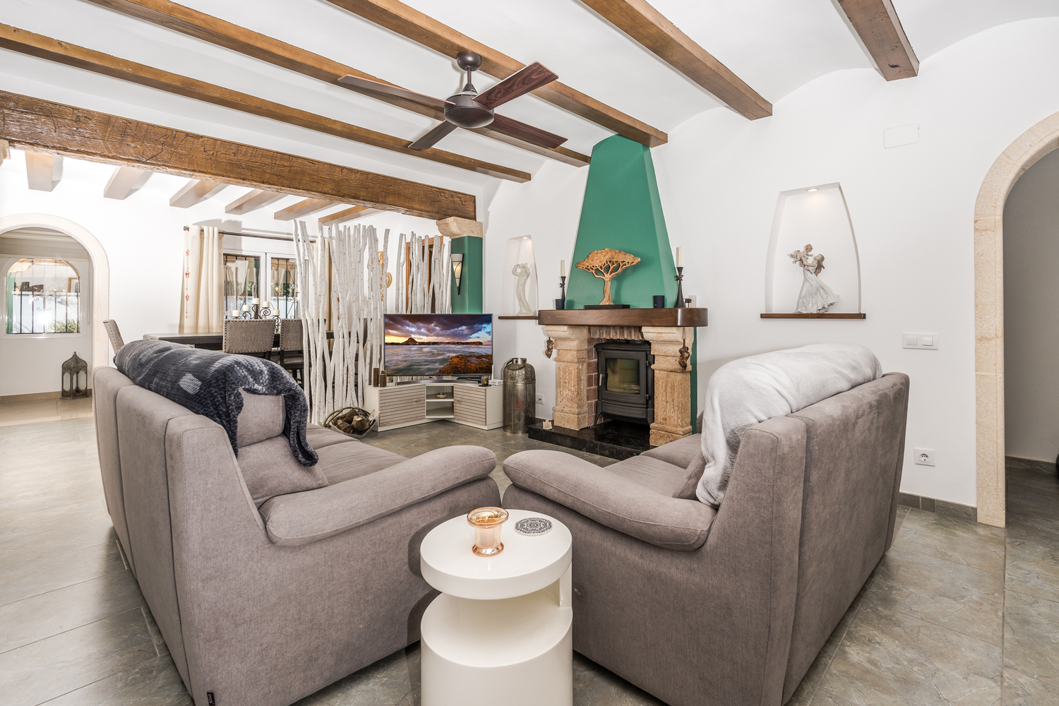 Living room featuring exposed wooden ceiling beams, a rustic stone fireplace with wood-burning stove, and tiled flooring.