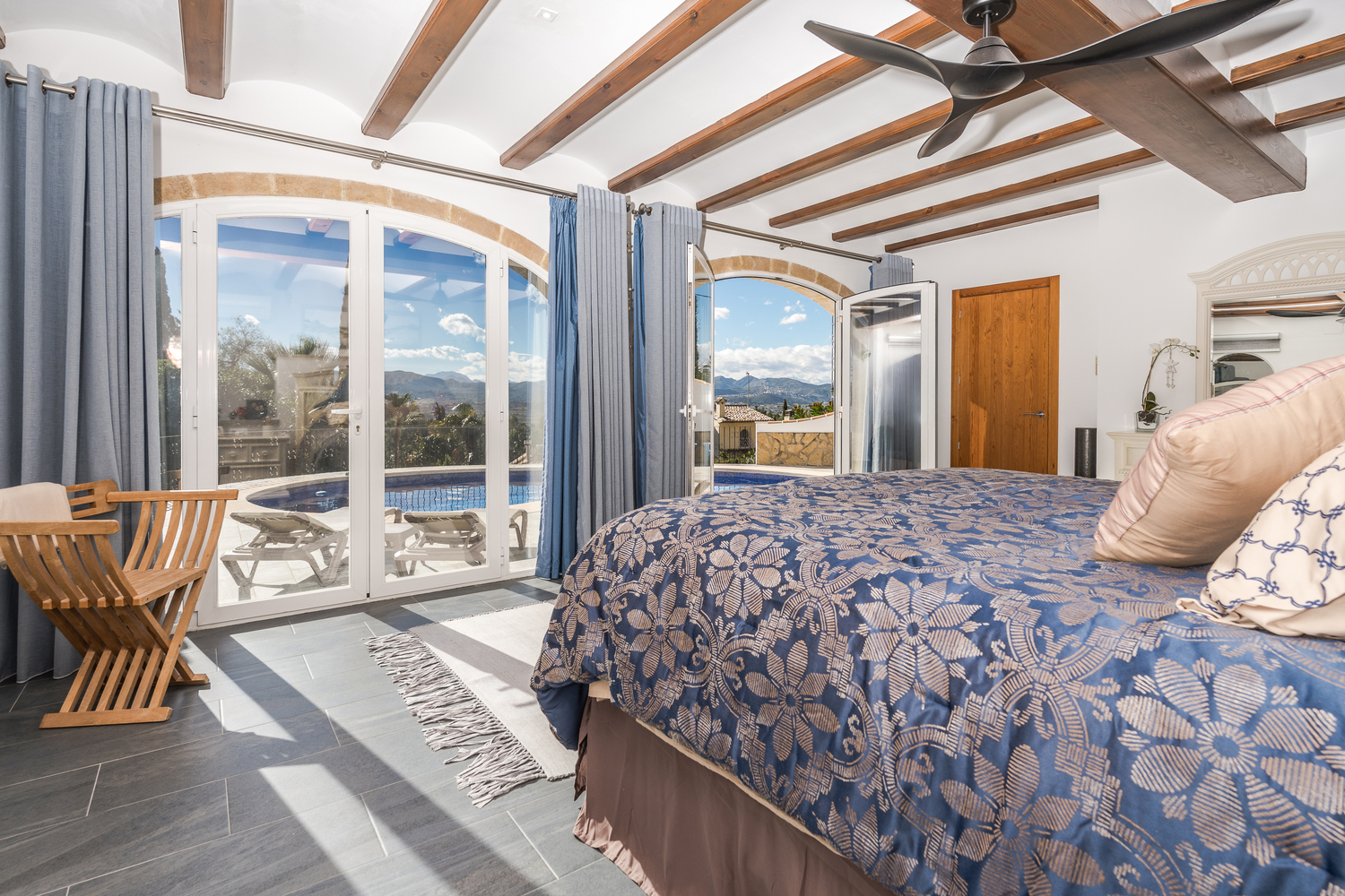 Master bedroom with exposed wooden beams, stone-arched glass doors offering direct pool access and panoramic valley views.