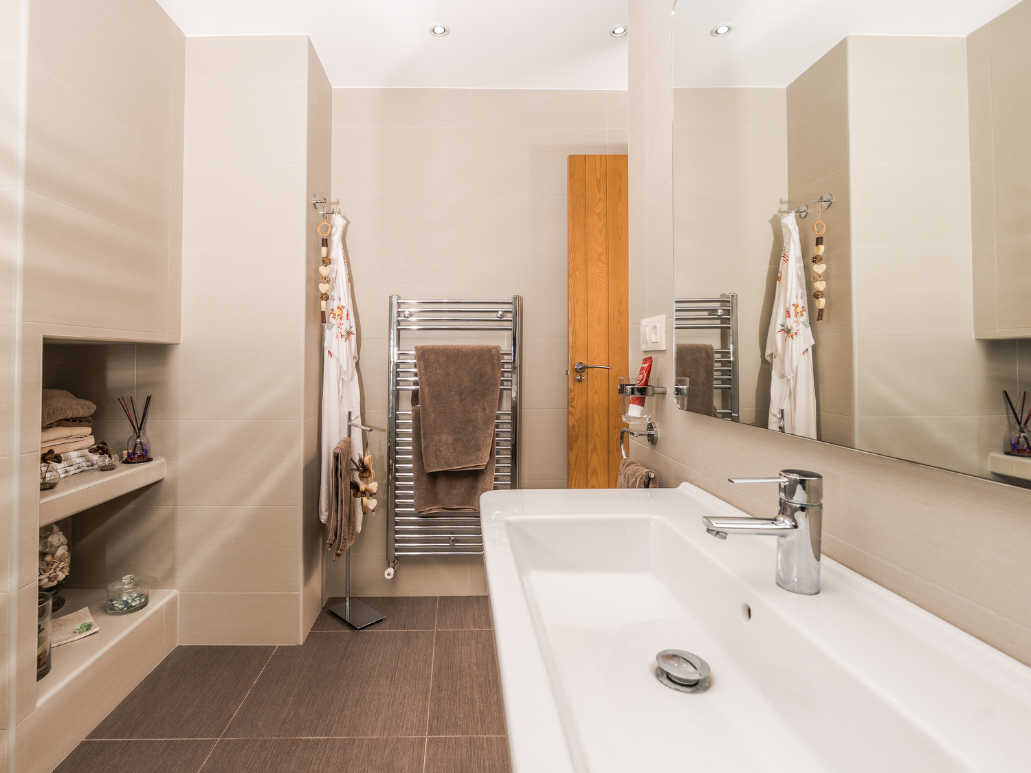 Modern bathroom featuring a large ceramic washbasin, recessed wall shelving, and a chrome heated towel rail against neutral beige tiling.