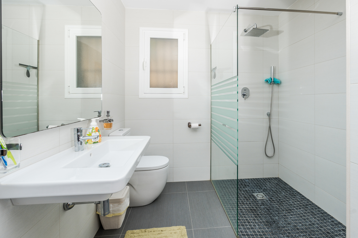 Modern bathroom featuring a walk-in shower with glass partition, dark mosaic flooring, and a wall-mounted sink. A frosted window provides natural light against white tiling.