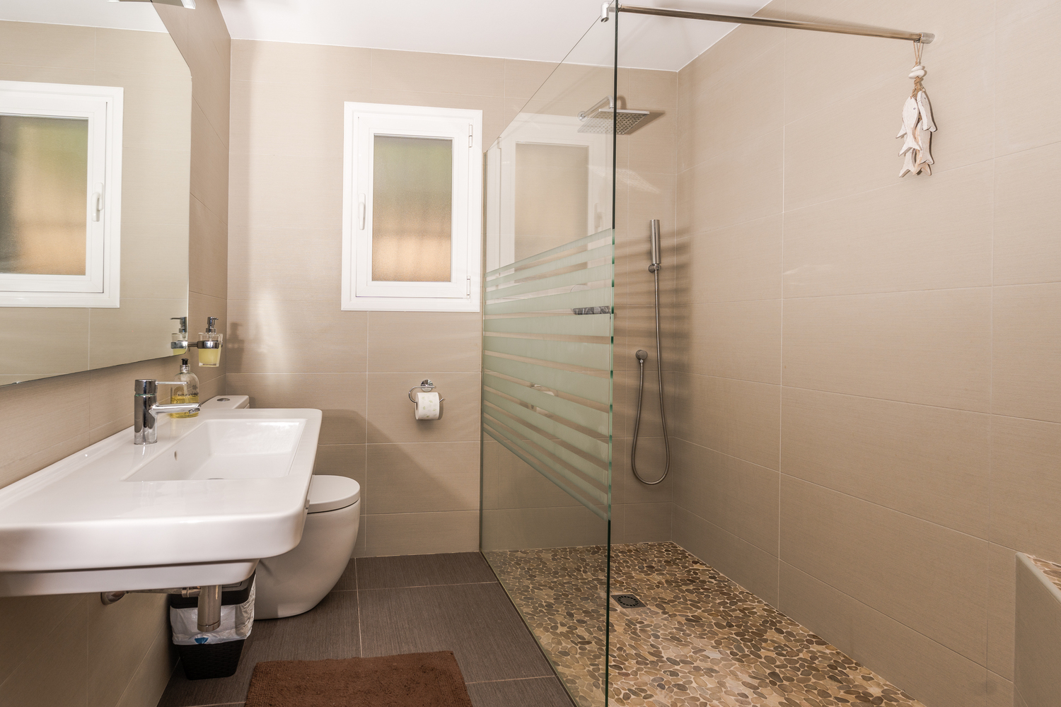 Modern bathroom featuring a walk-in shower with pebble flooring, a glass partition, and neutral-toned tiling. Includes a wall-mounted sink and frosted window.