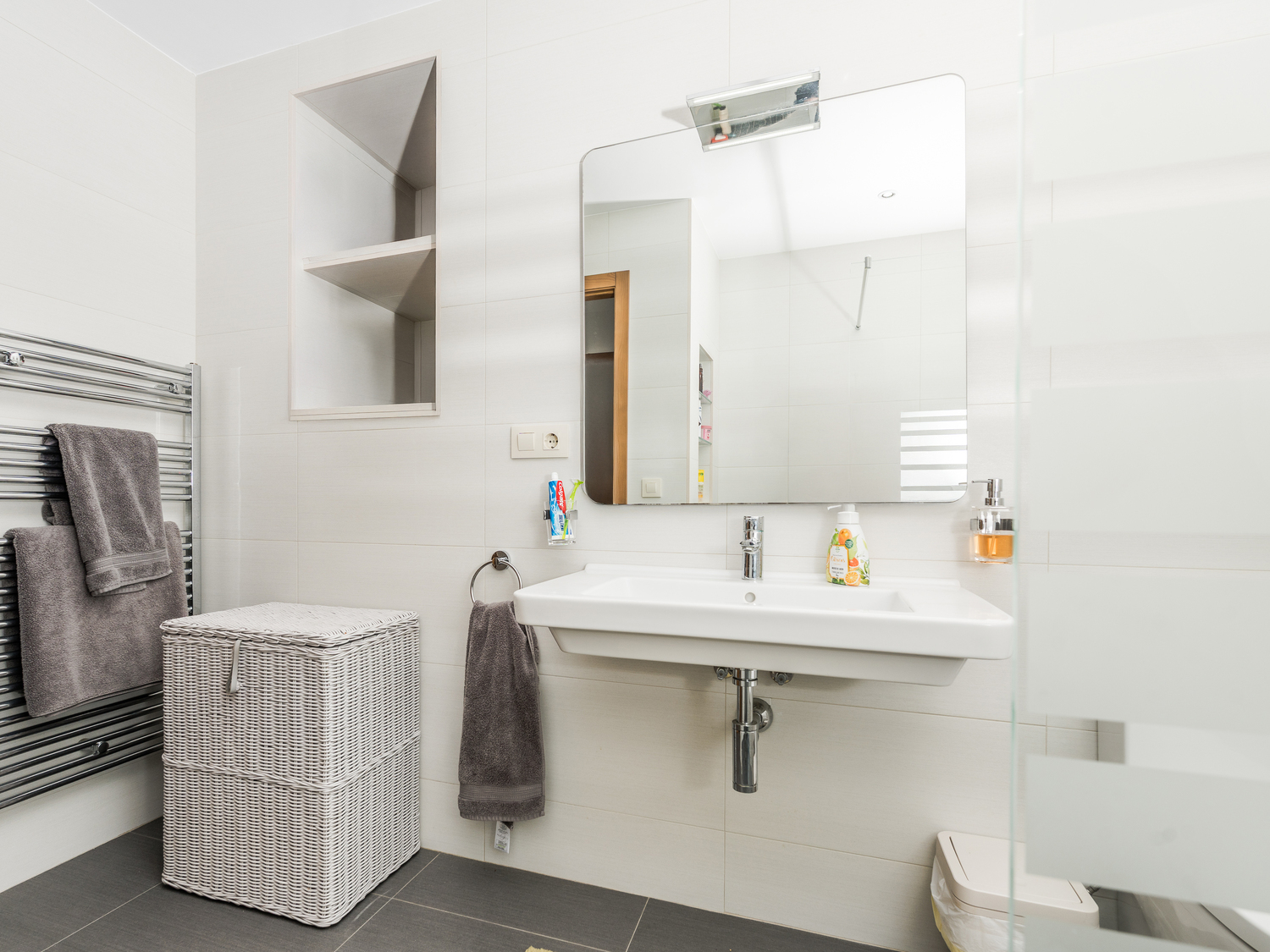 Modern bathroom featuring a wall-mounted ceramic sink, built-in storage niche, and chrome heated towel rail against white tiled walls.
