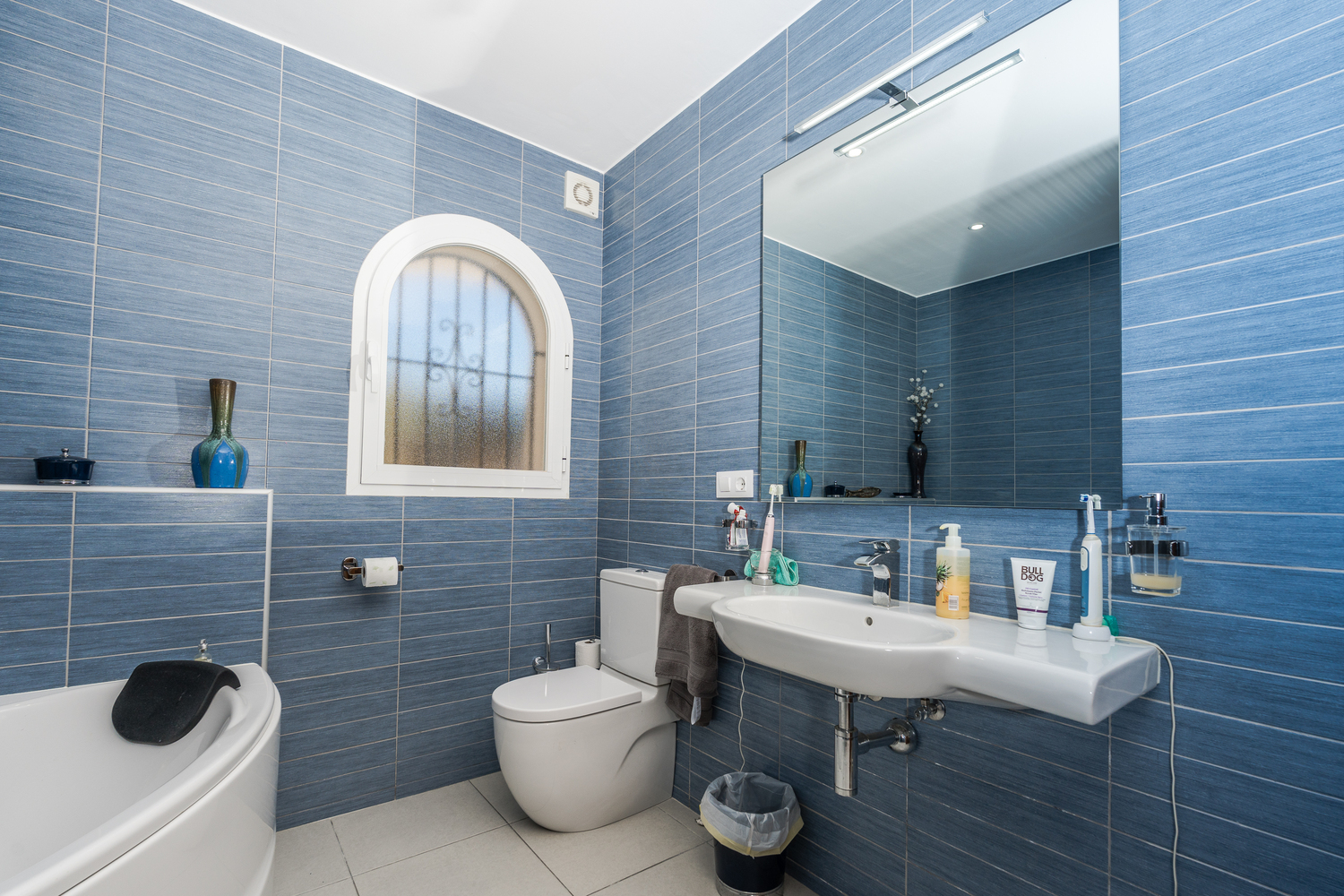 Modern bathroom featuring blue textured wall tiles, an arched window with privacy glass, a white ceramic sink, and a large mirror with integrated lighting.